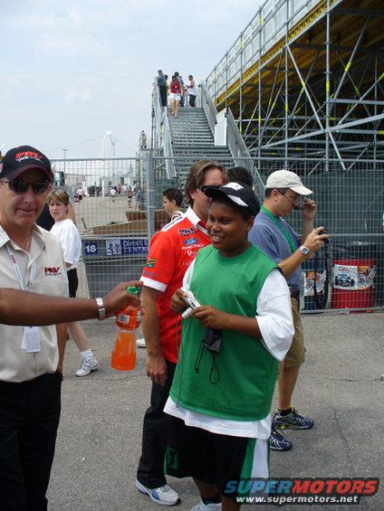 Man in PKV shirt hands drink to kid in green vest, race track stands in background