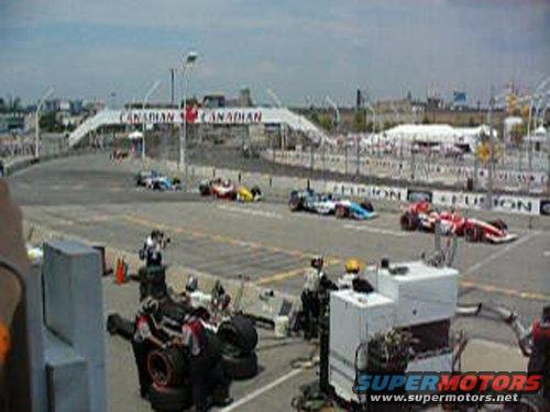 Race cars on a track with pit crew and Canadian banners overhead