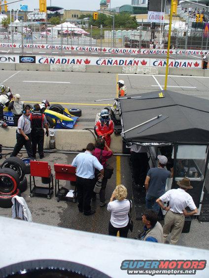 Race car pit crew and media gathered around a car in a pit stop area. A driver in a red suit stands near a tent