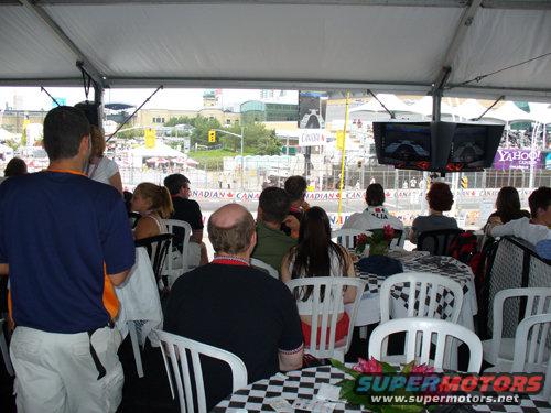 Spectators seated at checkered-clothed tables under a tent, watching a race on screens