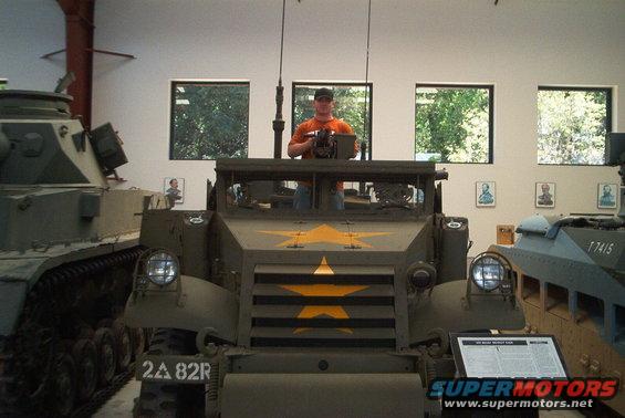 Front view of olive drab armored vehicle with yellow stars, man behind machine gun, other military vehicles nearby