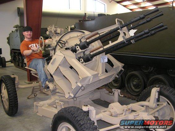 Person sits in the operator's seat of a quad-barreled anti-aircraft gun mount. Another tracked military vehicle is visible behind it