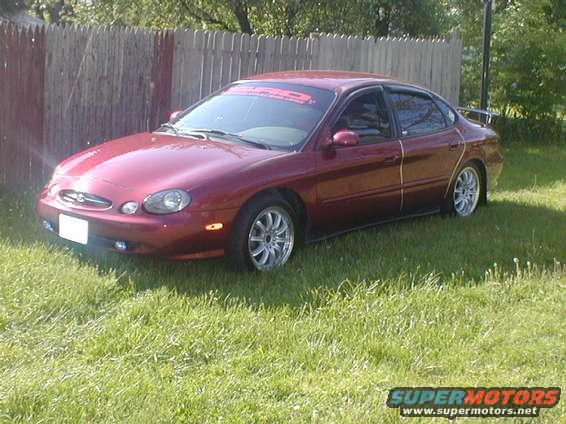 Maroon sedan parked in front of a wooden fence. It has aftermarket wheels and a banner on the windshield