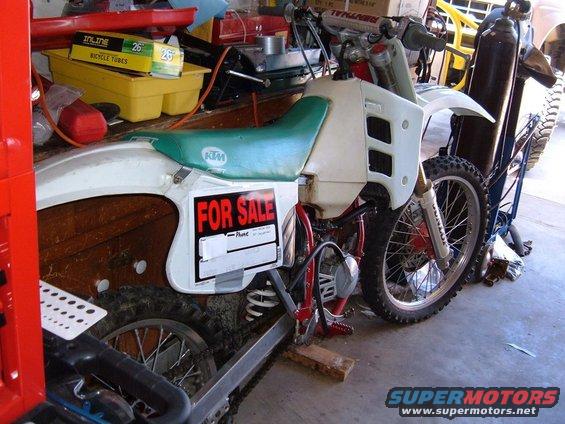 White dirt bike with a green seat and a "FOR SALE" sign on its side, resting on blocks in a garage