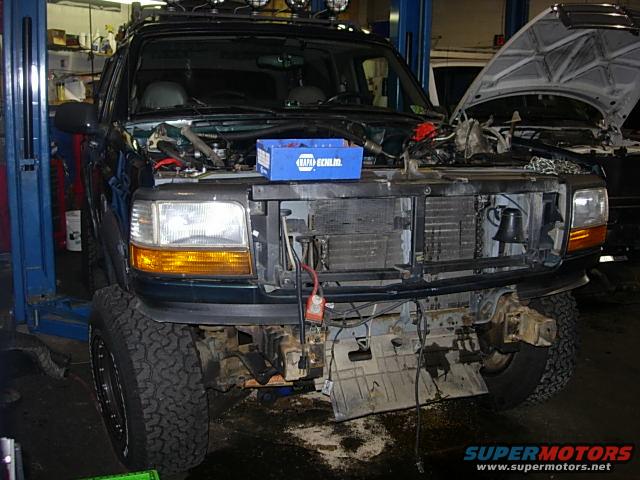 Front of dark green Ford F-Series truck with hood open, engine exposed, and front bumper removed. It's on a lift in a garage