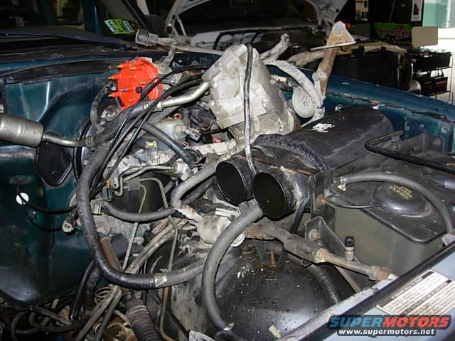 Engine compartment with distributor, hoses, and air intake visible. Supermotors.net logo in lower right