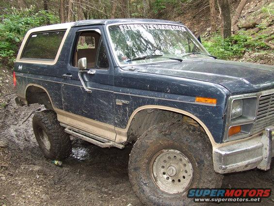 Dark blue Ford Bronco with a white top, lifted with large tires, driving through mud. Text on windshield reads 'KUTZNERZEBROCKON'