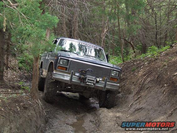 Ford Bronco driving through a muddy, wooded trail. Windshield banner says "BRONCO.COM"