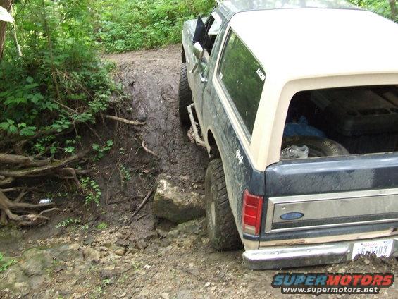 Ford Bronco with a tan top on a muddy, wooded trail, rear view