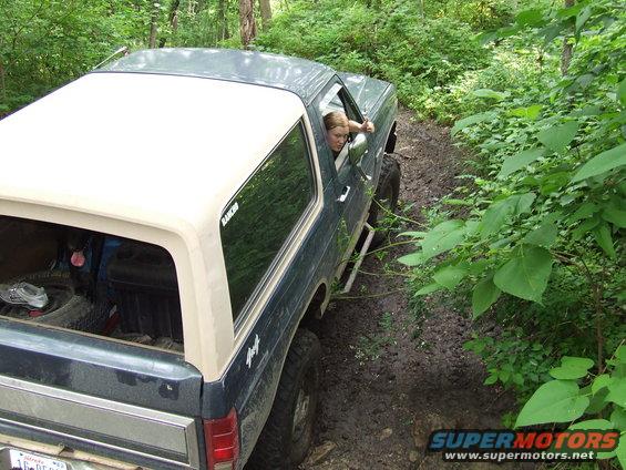 Dark green Ford Bronco with a white top driving on a muddy trail in the woods. A woman is in the driver's seat