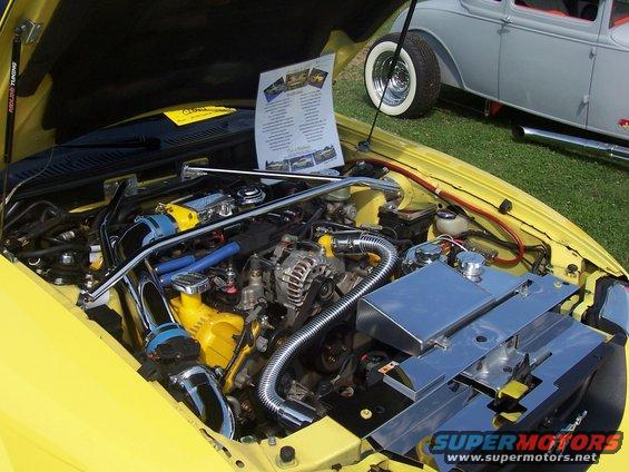 2002 Ford Mustang engine bay with chrome intake and yellow engine components. Another car is visible in the background