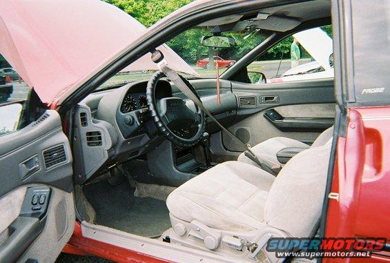 red car interior, seats, steering wheel, dashboard, and door panel visible. Another red car and a white car are in the background