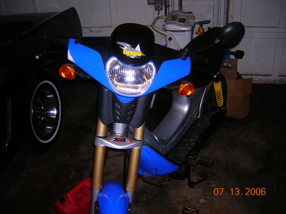 Blue and black scooter with gold front forks and a yellow shock absorber, parked indoors next to a black car