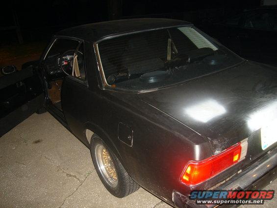 Black coupe with gold mesh wheels, driver's door open, showing tan interior. Rear taillight visible