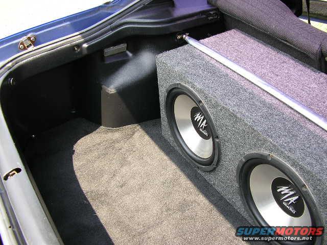Car trunk interior showing a carpeted subwoofer box with two MA audio speakers and a metal strut bar