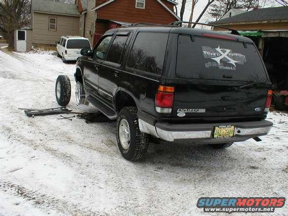 Black Ford Explorer SUV, rear view, jacked up with a wheel removed, snow on ground