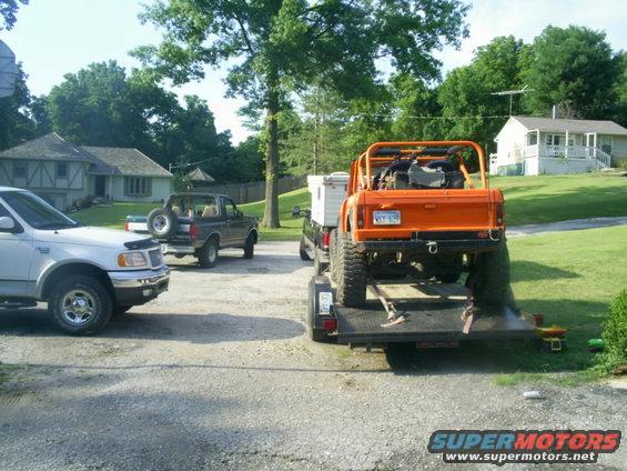 Orange off-road vehicle on a trailer, with a white pickup truck and a dark SUV in the background