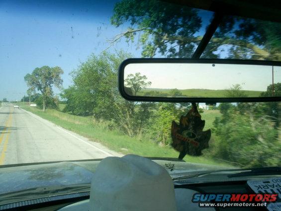 Driving down a country road, looking out the windshield. A cowboy hat is on the dashboard. Rearview mirror shows trees and fields. Another car is visible on the road ahead