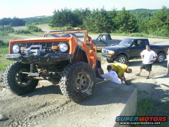 Orange off-road vehicle with winch and large tires, parked on dirt. Two Ford trucks are behind it