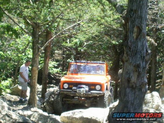 Orange off-road vehicle with large tires navigating a rocky trail, a man stands nearby
