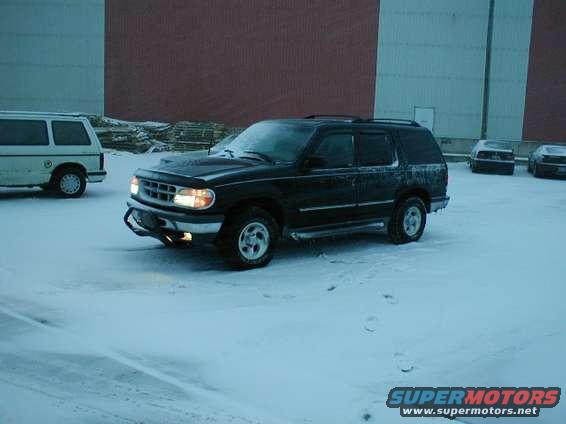 Black Ford Explorer parked in snow, front end visible. White van to the left, other cars in background