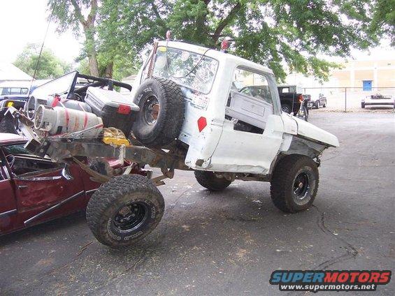 White pickup truck chassis with spare tire mounted on rear, propane tank and other items loaded. Another vehicle is partially visible underneath