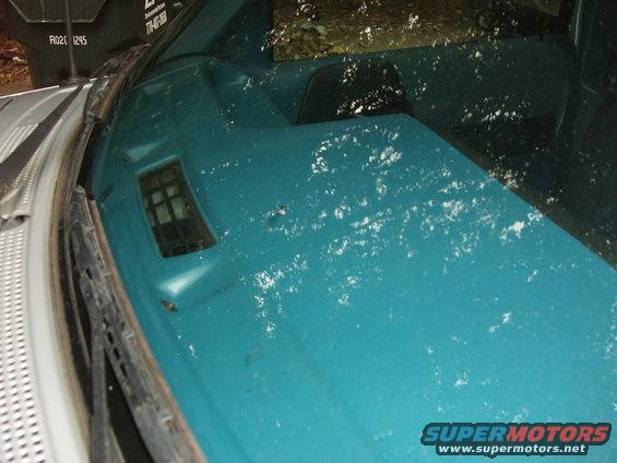 Close-up of a teal truck dashboard with white specks and a vent, showing part of the windshield and wiper. Text on a trash bin is visible