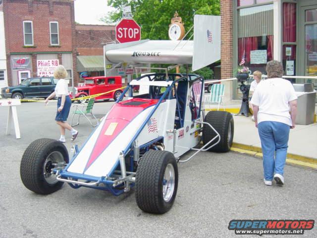 Open-wheel race car with Hoosier sponsorship and large rear wing, parked on street. Red car in background