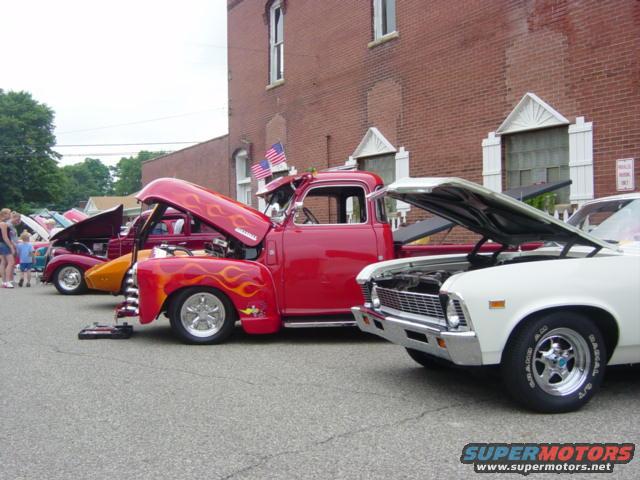 Red pickup truck with flames painted on hood and side, parked next to a white classic car. Both have hoods up