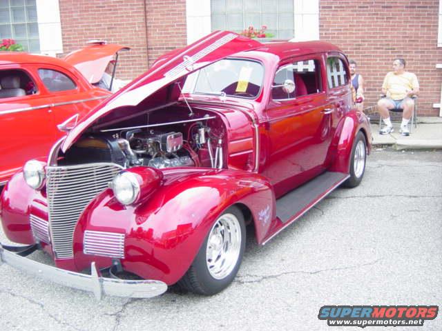 Red 1939 sedan with hood open, showing engine. Another red car is parked behind