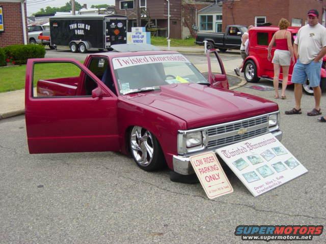 Red pickup truck with custom hood, open doors, and chrome wheels. Sign reads 'Low Rider Parking Only'