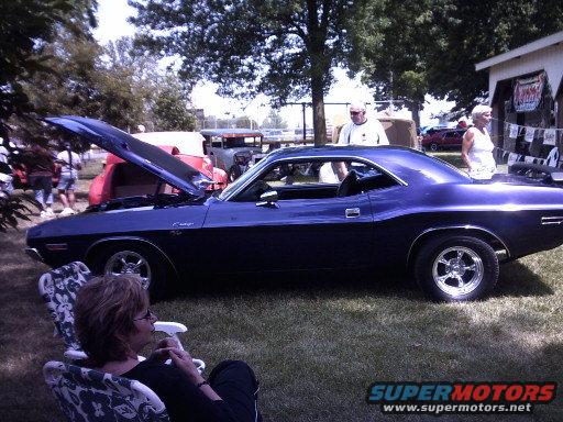 Dark blue muscle car, possibly a Dodge Challenger, with hood open parked on grass. A woman sits in a chair in the foreground