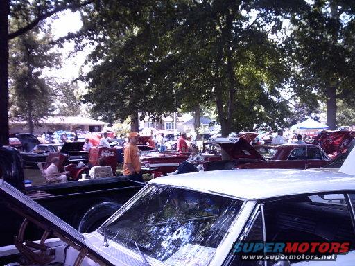 White car hood and windshield in foreground, with multiple classic cars and people in background under trees