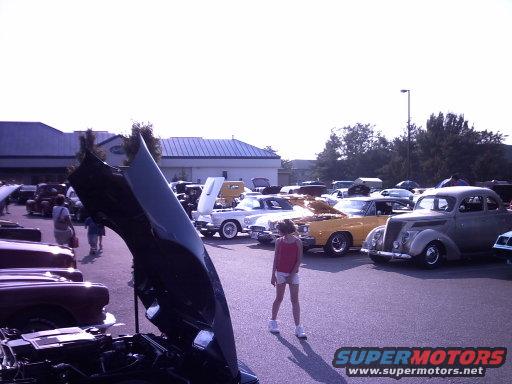 Car show scene with a young girl standing in front of several classic cars, one with its hood open