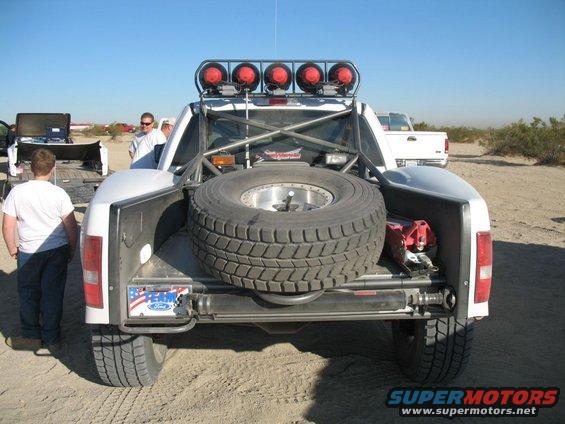 Rear view of a white off-road truck with a spare tire mounted in the bed, roll cage, and lights on top. Another white truck is visible in the background