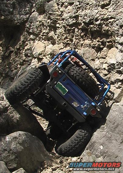 Blue off-road vehicle with roll cage tilted on rocks, showing spare tire and green license plate