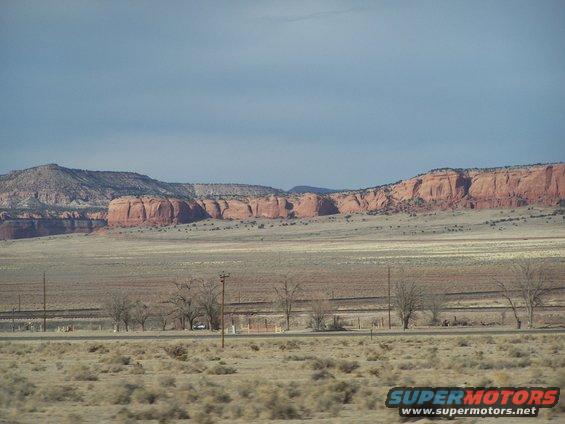 Desert landscape with red rock mesas under a cloudy sky, a road runs through the middle ground
