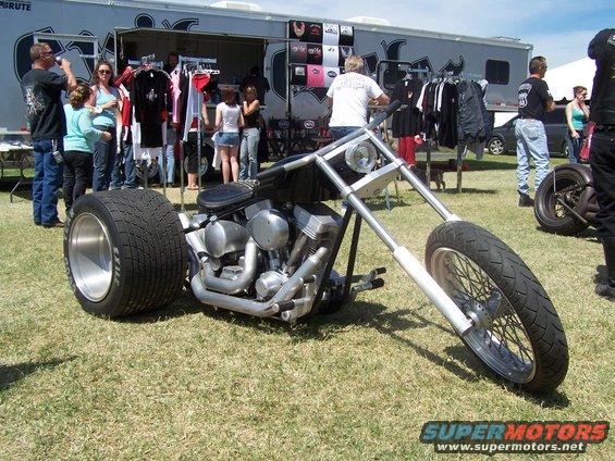 Chopper motorcycle with wide rear tire and exposed engine, parked on grass in front of a merchandise trailer
