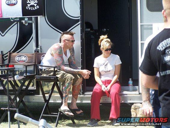 Man with tattoos and mohawk sits in a director's chair next to a woman in red pants sitting on steps outside a trailer