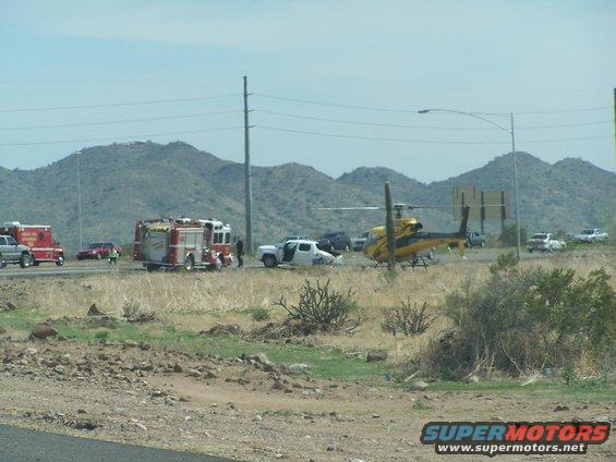 Emergency vehicles, including fire trucks and a helicopter, are at a scene on the side of a highway with mountains in the background