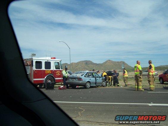 Fire truck and several people around a damaged blue car on the side of a highway