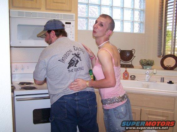Man in a blue cap and grey t-shirt with a motorcycle graphic stands at a stove, with another person in a pink and white striped top and denim skirt behind him