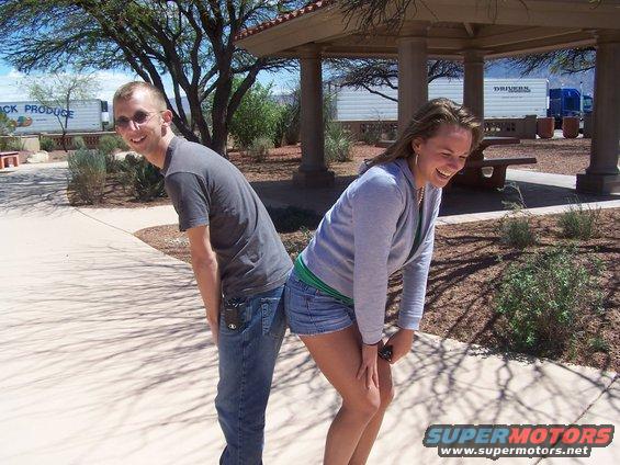 Man and woman bending over, backs to each other, laughing. Trucks in background