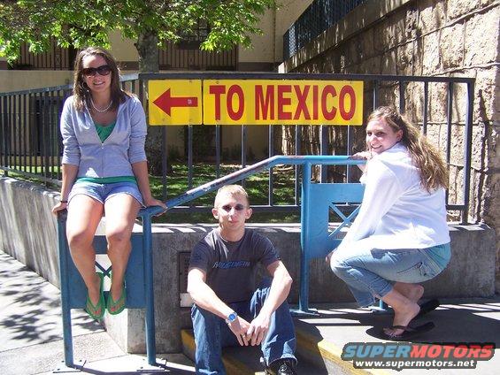 Three young people pose by a yellow sign pointing left with the words 'TO MEXICO'. One person sits on a railing, another squats, and one sits on steps