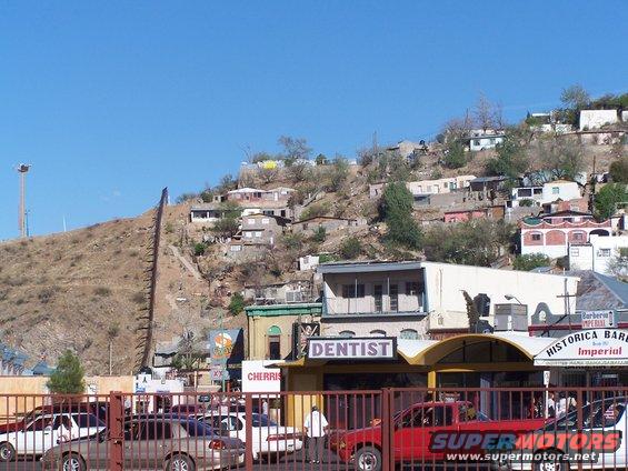 Border wall ascending a dry hillside towards a town of houses, with businesses and parked cars in the foreground