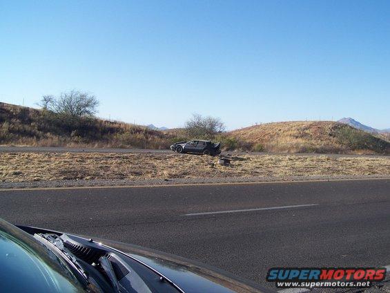 Dark car with a spoiler parked on the side of a highway, with dry grass and hills in the background