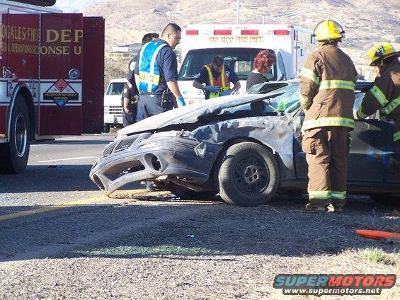 Car wreck with firefighters and police at the scene. The front of a dark sedan is heavily damaged