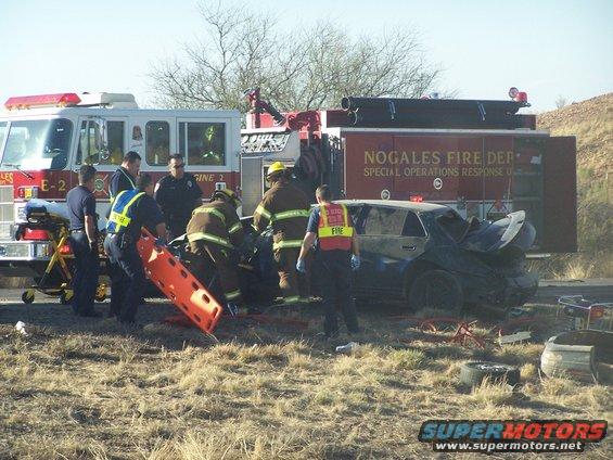 Damaged dark sedan at a crash scene with firefighters and police officers, a fire truck in the background