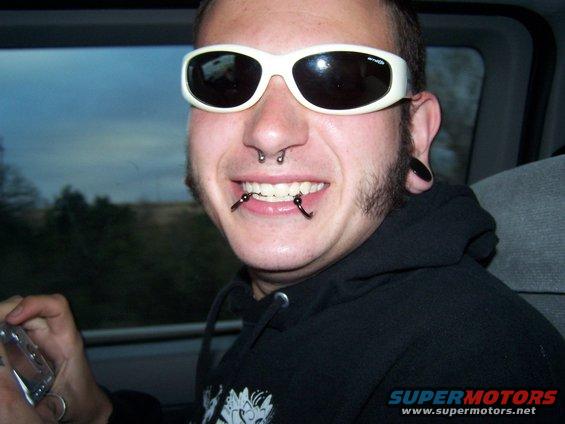 Man with white sunglasses and facial piercings smiles, holding a silver device