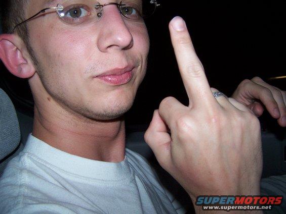 Man in glasses giving the middle finger, wearing a white t-shirt. Watermark reads SUPERMOTORS